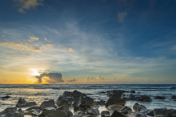 This image depicts a tranquil sunset scene along a rocky coastal shoreline, where the sun lowers toward the horizon, painting the sky in warm oranges, pinks, and golds amid scattered clouds. The foreground features large, weathered gray rocks partially submerged in shallow, glassy turquoise waters. In the background, a large cargo ship sails on the distant calm ocean under the fading light. The composition emphasizes natural serenity and vastness, with no people present. Suitable for commercial applications such as travel magazine covers, environmental reports on coastal ecosystems, tourism promotions for tropical destinations, book illustrations on marine geography, or website backgrounds for ocean-related businesses.