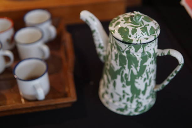 Enamel teapot with a distinctive green and white mottled pattern stands prominently next to blurred white cups on a wooden tray