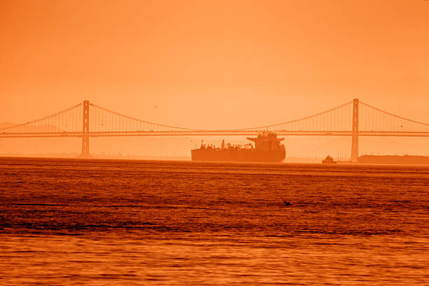 Orange Sunset Silhouette Usa Bridge Ship Crossing Bay With Cargo Containers On Deck, Hazy Horizon And Warm