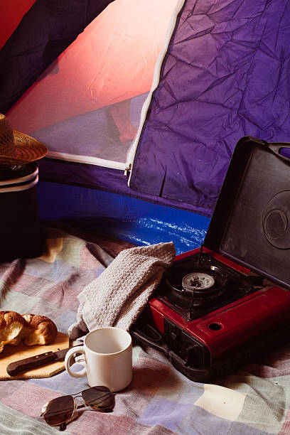 Camping breakfast scene with a portable stove, mug, croissant, and sunglasses laid on a checkered blanket outside a partly opened tent, enjoying morning outdoors leisure time