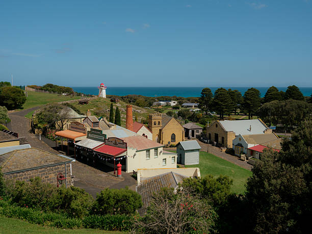 An elevated view looks down on an old  port town in Australia. The heritage village  overlooks the  deep blue ocean stretches across the horizon under a clear sky, framing the historic settlement nestled between lush green trees and coastal bluffs