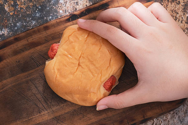 Close-up image shows a hand girl holding a sausage bun placed on a wooden cutting board.