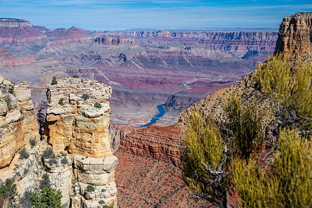 A majestic midday view over the Grand Canyon reveals a vast expanse of layered rock formations in shades of red, orange, and beige. The intricate erosion patterns and dramatic depth showcase millions of years of geological history under a clear blue sky. A timeless symbol of the American Southwest.