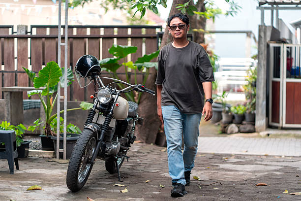 Asian Indonesian man wearing casual clothes walks beside a parked classic motorcycle in a quiet home yard. Lifestyle, transportation, freedom, and everyday urban living concept.