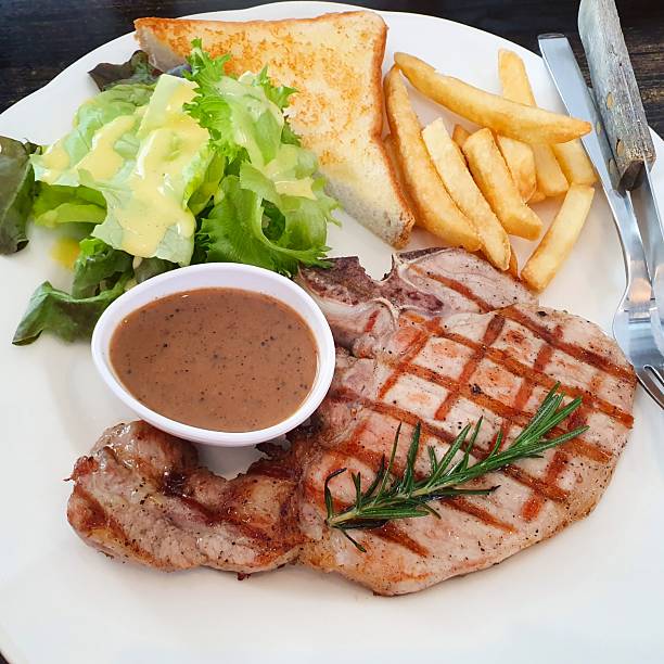 Delicious grilled pork chop steak with black pepper sauce served with toasted bread french fries and fresh garden salad on white plate