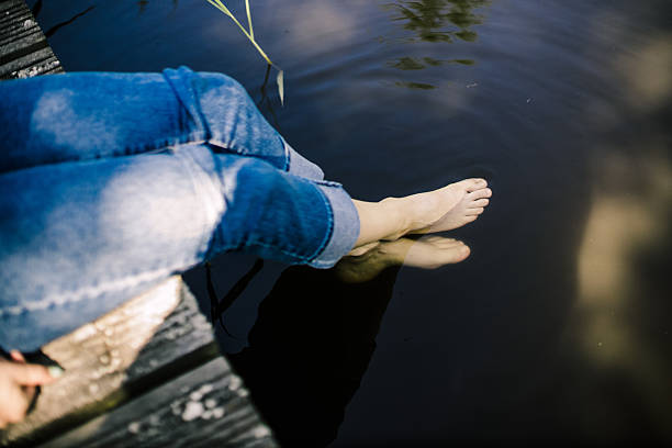 Close-up of bare feet gently touching calm lake water, creating ripples. Sunlight and shadows play on the serene surface, reflecting nature's tranquility and simplicity in a summer moment