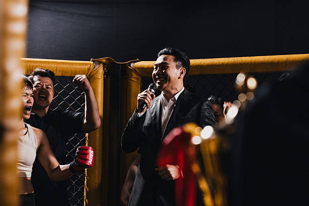 MMA announcer speaks into a microphone during a fight event in a cage setting with supporters cheering in the background.