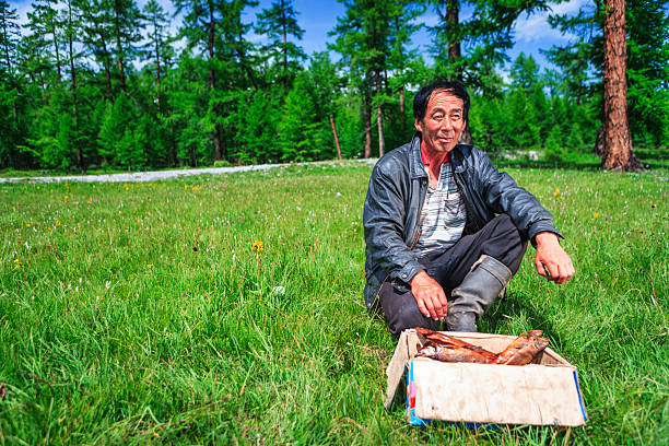 A middle-aged man sits cross-legged on a lush green meadow bordered by tall pine trees and a bright blue sky. He wears a worn leather jacket, striped shirt and rubber boots, and beside him is a cardboard box filled with smoked fish. The scene feels peaceful and rural, with sunlight, wildflowers and forested hills framing a quiet moment after a day of fishing.