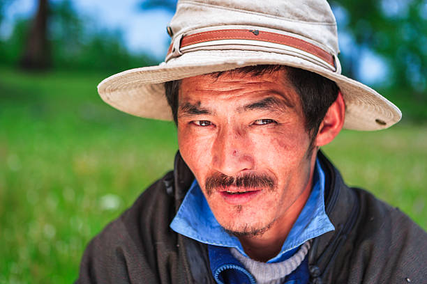 Close-up color portrait of a middle-aged man wearing a wide-brimmed sun hat and layered clothing. He has a mustache and short beard, slightly squinting eyes, and a weathered, sun-kissed complexion. The soft-focus green meadow in the background creates a calm, natural outdoor atmosphere.