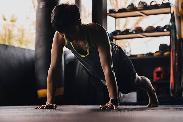 Female does push-ups in a gym during the day in a space with exercise equipment and sunlight coming through the windows.