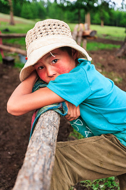 A young child with flushed cheeks and a woven sun hat leans on a rough wooden rail, clutching a colorful scarf. Dressed in a bright turquoise shirt and corduroy pants, the child gazes toward the camera against a blurred green, rural backdrop.