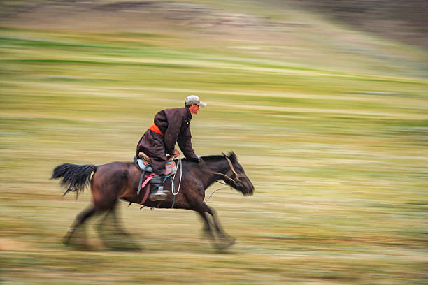 A dynamic panning shot of a rider leaning forward on a galloping horse across open grassy plains. Motion blur streaks the background into green and brown bands, emphasizing speed and movement; the rider wears a dark coat with a bright orange sash and a cap, and the horse is captured mid-stride with tack and flowing tail visible. The image conveys energy, traditional horsemanship, and wide, wind-swept landscape.