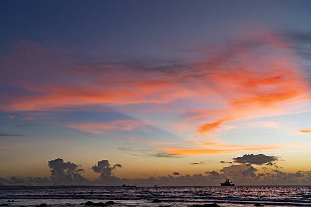 A small wooden boat drifts gently on the still surface of the ocean during sunset, silhouetted against a dramatic sky transitioning from deep blue to warm orange and pink hues. Scattered clouds enhance the colorful display over the horizon, with gentle waves lapping at rocky shores in the foreground. The scene captures a peaceful maritime moment, ideal for commercial applications such as travel brochures promoting coastal destinations, wellness campaigns featuring nature escapes, book covers for adventure novels, website headers for tourism sites, or editorial content on environmental conservation and ocean themes.