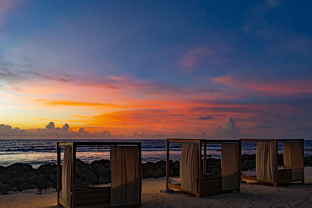 Four wooden beach cabanas, each draped with light white curtains, stand in a row on a sandy beach edged with rocks. The scene faces the calm ocean waves under a dramatic sunset sky blending orange, pink, purple, and blue hues. The tropical setting evokes tranquility and luxury relaxation. This image suits commercial uses in travel magazines, resort brochures, lifestyle blogs, and promotional materials for vacation destinations, highlighting serene coastal escapes and sunset vistas for editorial and advertising content.