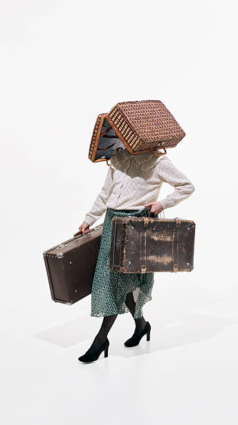 Woman in retro outfit carrying old suitcases with wicker basket covering head on white background. Concept of relocation, identity crisis, travel burden, moving house and life transition.