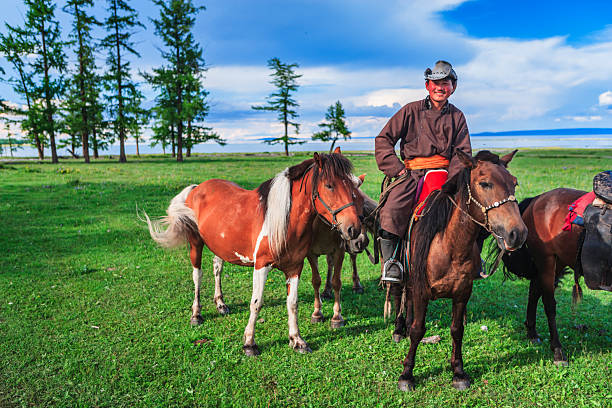 A smiling rider in traditional attire sits on a brown horse while two other horses, including a chestnut-and-white pony, stand close by on a lush green meadow. Scattered tall trees frame a calm lake in the background beneath a bright blue sky with soft clouds. The sunlit scene conveys a peaceful, pastoral moment with vivid colors and relaxed animals.