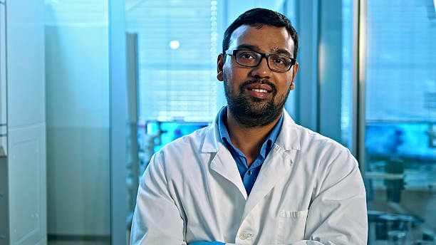 A South Asian male scientist in a white lab coat and glasses stands confidently in a modern lab, surrounded by advanced equipment, highlighting medical research and healthcare innovation.
