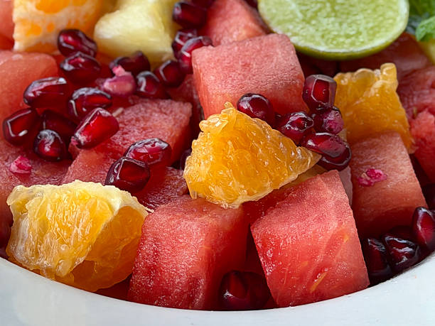 Full frame image of fruit salad in white bowl, cubed watermelon, diced orange, pomegranate seeds, pineapple pieces, lemon slice and mint leaf garnish, wood grain kitchen counter, elevated view