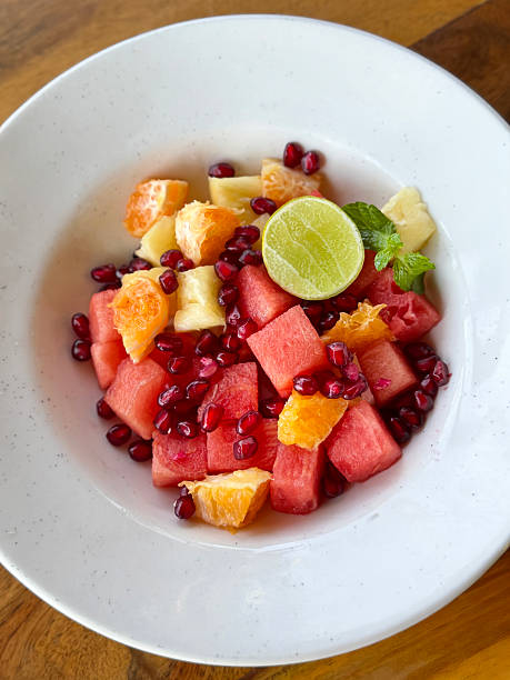 Image of fruit salad in white bowl, cubed watermelon, diced orange, pomegranate seeds, pineapple pieces, lemon slice and mint leaf garnish, wood grain kitchen counter, elevated view