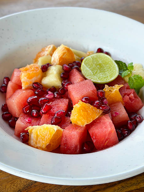 Close-up image of fruit salad in white bowl, cubed watermelon, diced orange, pomegranate seeds, pineapple pieces, lemon slice and mint leaf garnish, wood grain kitchen counter, elevated view