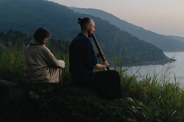 Man playing wind instrument beside woman on coastal cliff.