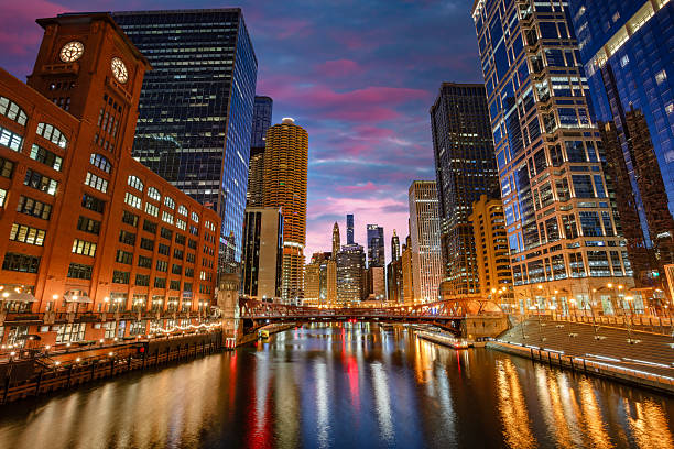 Chicago, Illinois, USA. The downtown skyline is captured at sunrise, featuring tall skyscrapers on either side of the river. The sky displays beautiful pink and purple hues. Prominent buildings, including those with large glass facades, reflect lights in the water. The river is calm, reflecting the city's lights. A bridge in the middle distance adds depth to the image. The scene is illuminated by streetlights, enhancing the urban atmosphere in the early morning light.
