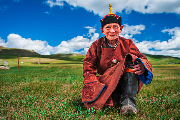 An elderly man kneels on a grassy steppe, smiling gently at the camera. He wears a richly embroidered rust-red robe with blue lining and decorative fastenings, and a tall, colorful hat topped with a golden ornament. His weathered hands rest on a sturdy, ornate boot; deep lines on his face suggest a life spent outdoors. The scene is set against rolling green hills under a vivid blue sky dotted with white clouds, with warm sunlight bringing out the textures of his clothing and the grass.