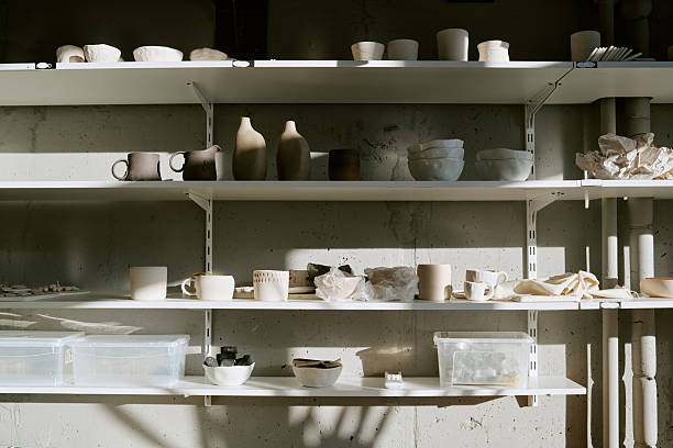 Ceramic bowls, cups, vases and pottery pieces arranged on shelves in studio setting, sunlight casting shadows on wall, various handmade objects displayed in organized manner