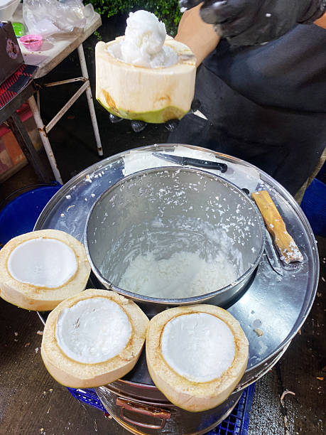 Coconut ice cream being served in a coconut shell, with multiple empty coconut shells on a metal container, showcasing a street food preparation scene