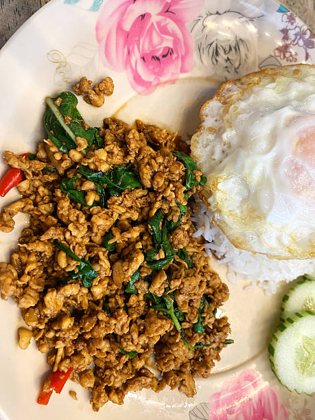 Stir-fried minced chicken with basil served on a plate alongside fried egg and cucumber slices, garnished with red chili and herbs