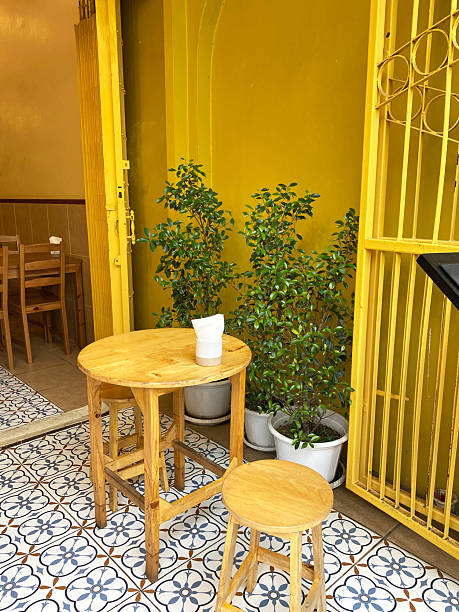 Wooden table and stool set outside a cafe with yellow walls and potted plants, featuring patterned tiles and an open door inviting customers to enter