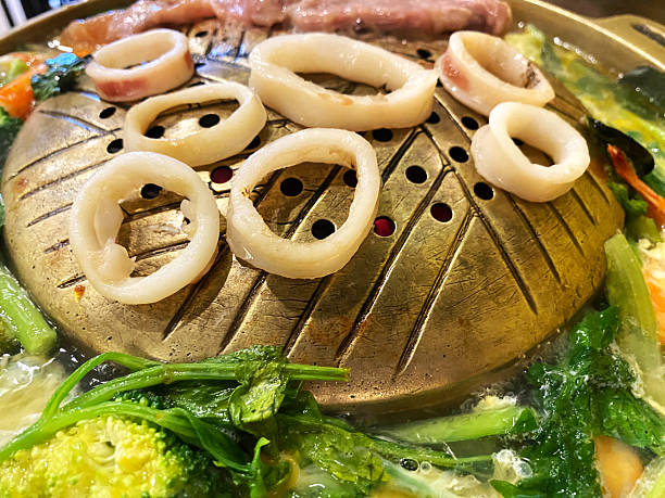 Hot pot cooking scene featuring fresh squid rings and assorted vegetables simmering in a metallic pot at a vibrant restaurant setting