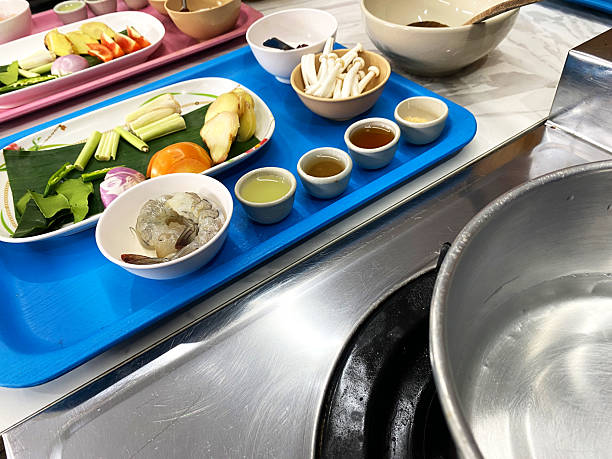 Ingredients for preparing Thai Tom Yum soup displayed on blue tray, including shrimp, herbs, vegetables, and sauces, with a pot of water in the background