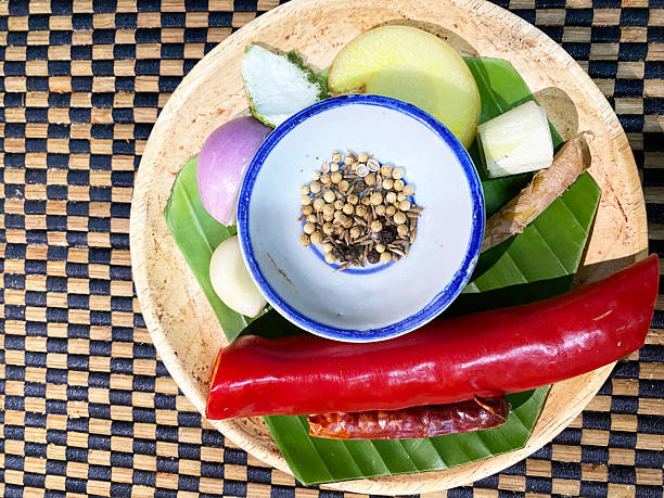 Assorted spices and fresh ingredients for preparing Thai cuisine arranged on a round wooden plate with green banana leaves and a woven mat background