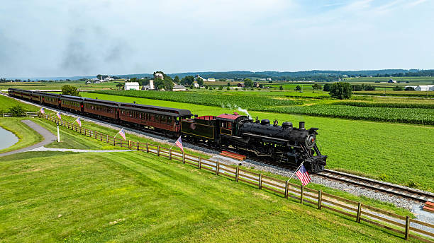 A steam train moves along the tracks in a rural area of Pennsylvania. The train has many cars and passes by green fields and distant farms. Flags are placed along the railway.