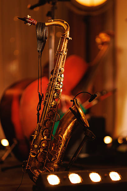 A close up of a brass saxophone placed on a stage stand. In the background, other orchestral instruments and warm stage lights are visible.