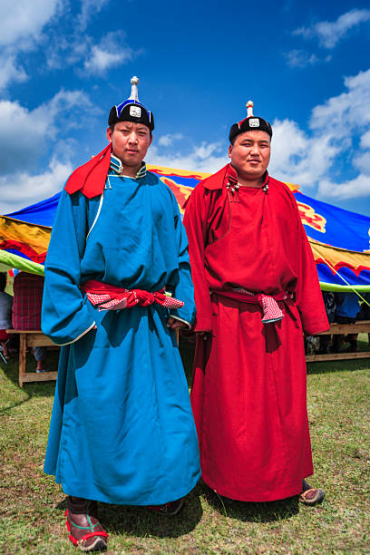 Two men stand side by side on grassy ground in front of a brightly colored festival tent under a vivid blue sky with scattered clouds. One wears a long blue robe and the other a long red robe, each cinched with patterned sashes and topped with traditional hats featuring small finials. The scene is sunlit and vibrant, capturing a calm, posed portrait at an outdoor cultural gathering.