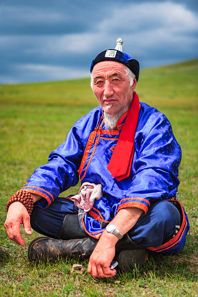 A seated elderly man on open grassland wears a bright blue, embroidered robe with orange trim and a red scarf, paired with a small traditional hat topped by a white ornament. He has a white goatee, weathered facial features and a calm, contemplative expression. Visible accessories include wooden prayer beads on his wrist, a wristwatch, sturdy leather boots, and a cloth tucked at his waist; the scene is set against rolling green hills under a moody, cloud-filled sky.