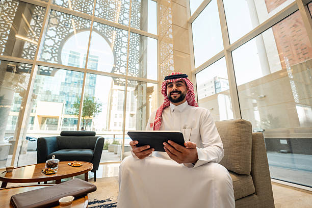 Middle Eastern man in modest clothing sits in a contemporary hotel lobby, working on a digital tablet. Natural light floods through expansive windows, illuminating the modern architectural space with glass and metal accents