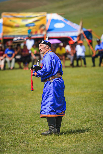 An older man stands on a grassy field wearing a bright blue silk deel (traditional robe), a decorated belt and tall black boots. He holds a small ornate hat with red tassels and looks toward an off-frame activity. Behind him, colorful festival tents and a seated crowd are softly blurred, suggesting a lively outdoor cultural event on a sunny day.