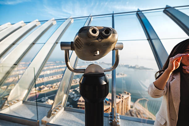 Binoculars and view of the Palm Jumeirah coastline from the observation deck. Structures of the Dubai skyline are visible through the transparent glass panels. The sunlight illuminates the city during the day. Leisure activity and exploration