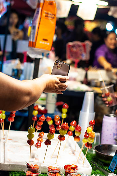Customer picking a strawberry tanghulu and paying by electronic money transfer via mobile phone at a night market stall at Thai street food market at Bangkok Thailand Night Market digitally generated image stock photo copy space vertical