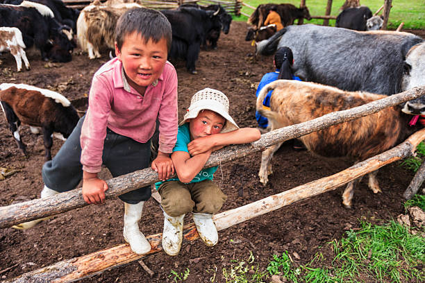 Two young children lean on a simple wooden fence inside a muddy livestock pen, surrounded by yaks and goats. One child in a pink shirt smiles toward the camera while the other, wearing a sun hat and white rubber boots, sits on the lower rail with a more reserved expression. The image captures a candid moment of daily pastoral life, with animals, worn boots, and earthen ground filling the frame.