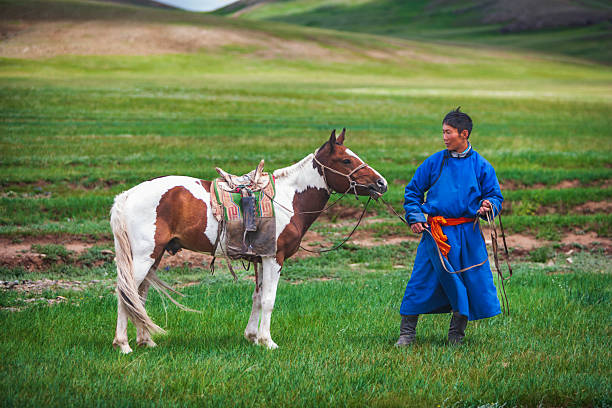 A person dressed in a bright blue traditional robe with an orange sash stands on a lush green steppe, holding the reins of a saddled pinto horse. The horse is white and brown with a colorful saddle blanket, and both figures are framed by gently rolling grassy hills under soft daylight. The scene conveys a quiet pastoral moment in an open rural landscape.