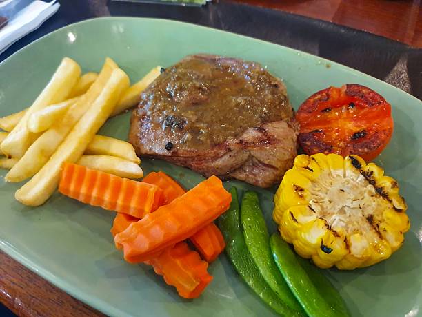 Delicious Grilled Beef Steak Topped with Savory Black Pepper Sauce Served with French Fries Roasted Corn and Fresh Vegetables on Light Green Plate