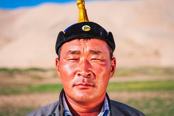 Close-up portrait of a middle-aged man facing the camera, wearing a black traditional cap topped with a small golden finial. Warm sunlight illuminates his weathered skin, faint stubble, and the lines around his eyes and forehead. His expression is calm and stoic. The background is softly blurred, showing pale sand dunes and hints of green, suggesting an open, arid landscape.
