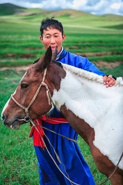 A smiling young person in a bright blue robe and red sash stands beside a brown-and-white pinto horse, one hand resting gently on the animal's neck. The horse wears a simple rope halter and reins; both figures are set against rolling green grasslands under a partly cloudy sky. The scene feels vivid and pastoral, emphasizing the close bond between rider and horse.