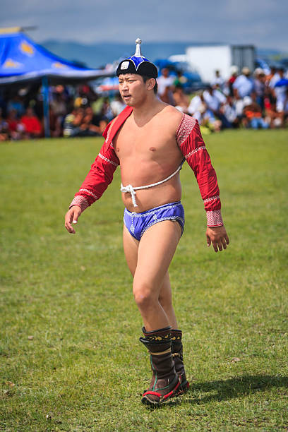 A young male traditional Mongolian wrestler walks across a grassy field wearing a red open-chested jacket, blue wrestling briefs and a white braided sash. He wears a pointed ceremonial hat and ornate leather boots, appearing focused as a blurred crowd watches an outdoor wrestling competition or festival in the background.