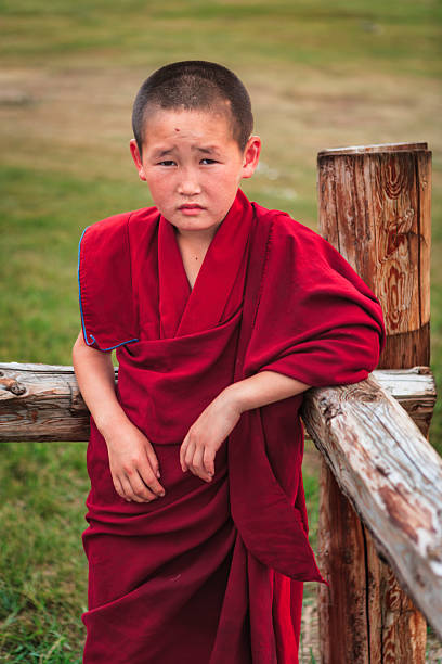 A young boy with a closely cropped haircut leans against a weathered wooden fence post in a grassy field. He wears deep red monastic-style robes draped over his shoulder and holds a quiet, slightly pensive expression. The background is softly blurred meadow, emphasizing the subject and the textured wood of the fence.