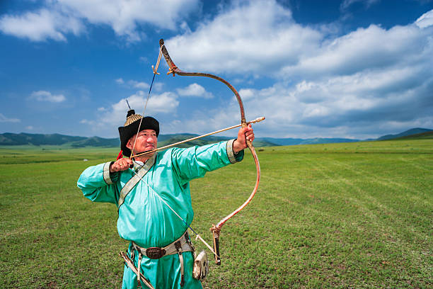 A man in bright turquoise traditional clothing and a fur hat draws a curved wooden bow, steadying an arrow as he aims across a wide green plain. Rolling hills and a dramatic blue sky with scattered clouds stretch into the distance, evoking open-space freedom and cultural tradition.